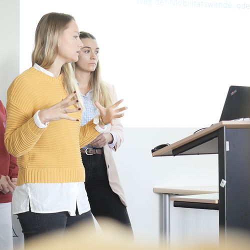 Auf dem Bild sind drei Studentinnen zu sehen, die vor einer Gruppe Menschen einen Vortrag hält. In Hintergrund an einer weißen Wand lässt sich eine Präsentation erahnen. 