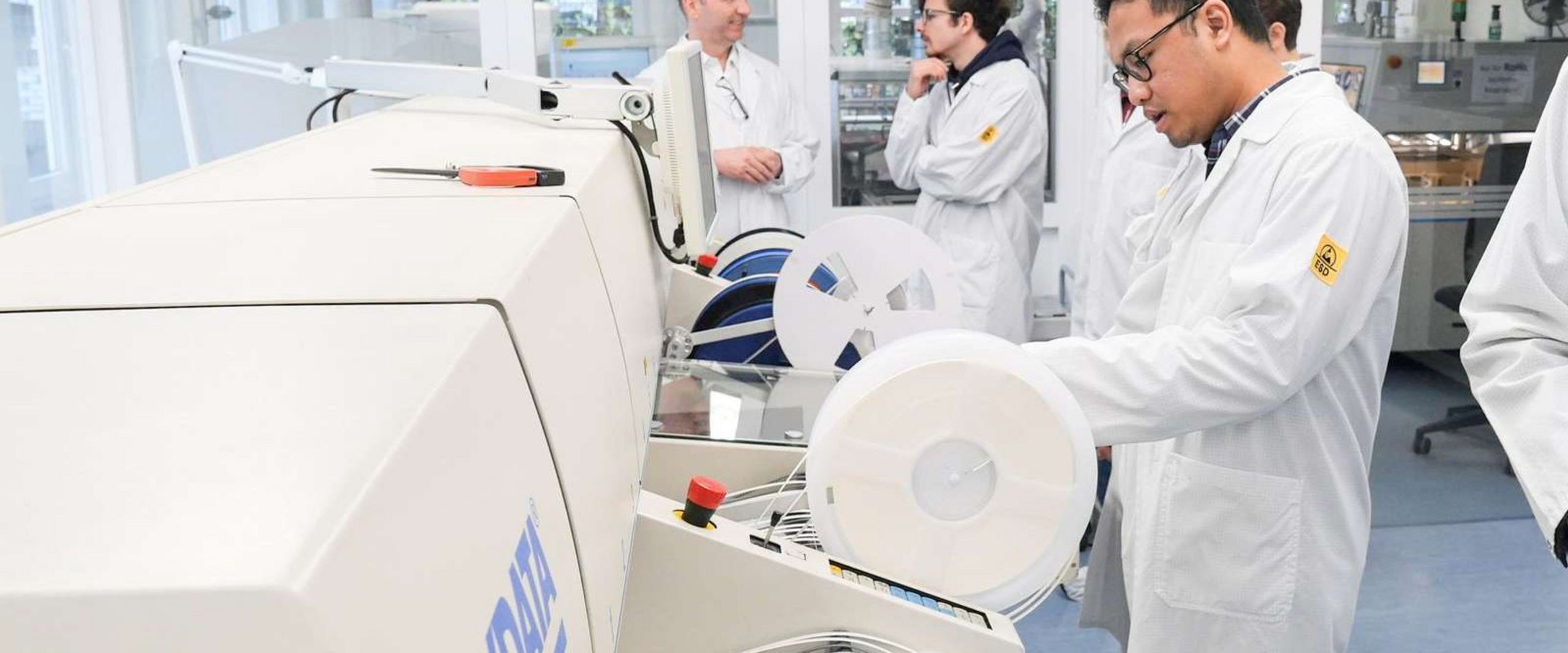 Students in lab coats working with electronic assembly equipment in a university laboratory.