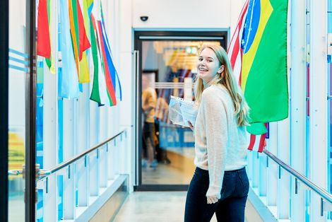 Studentin läuft einen Gang entlang, der von Flaggen gesäumt wird und blickt zurück in die Kamera.