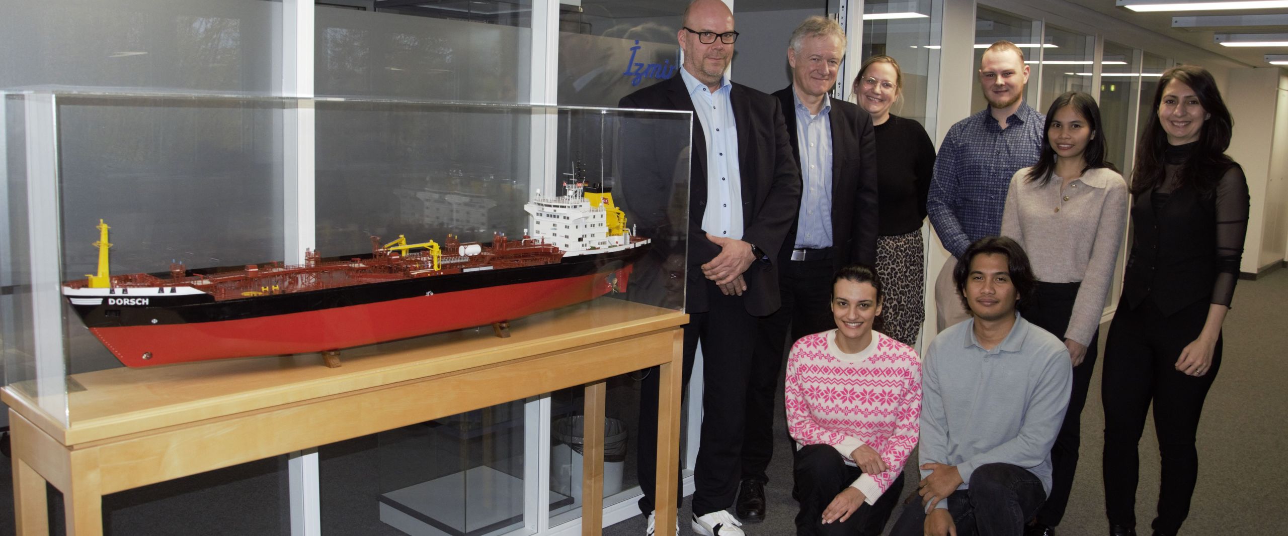 Gruppenbild mit Männern und Frauen. Links von ihnen steht ein Schiffsmodell in einer Vitrine.
