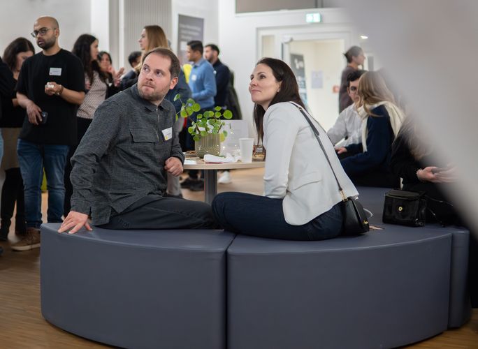 Eine Absolventin und ein Absolvent sitzen in einem Raum mit mehreren Leuten