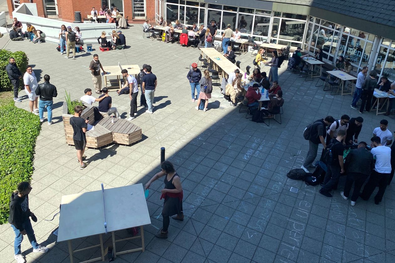Bild von oben auf Vorplatz von Geb&auml;ude. Menschen sitzen und stehen dort und spielen Tischtennis