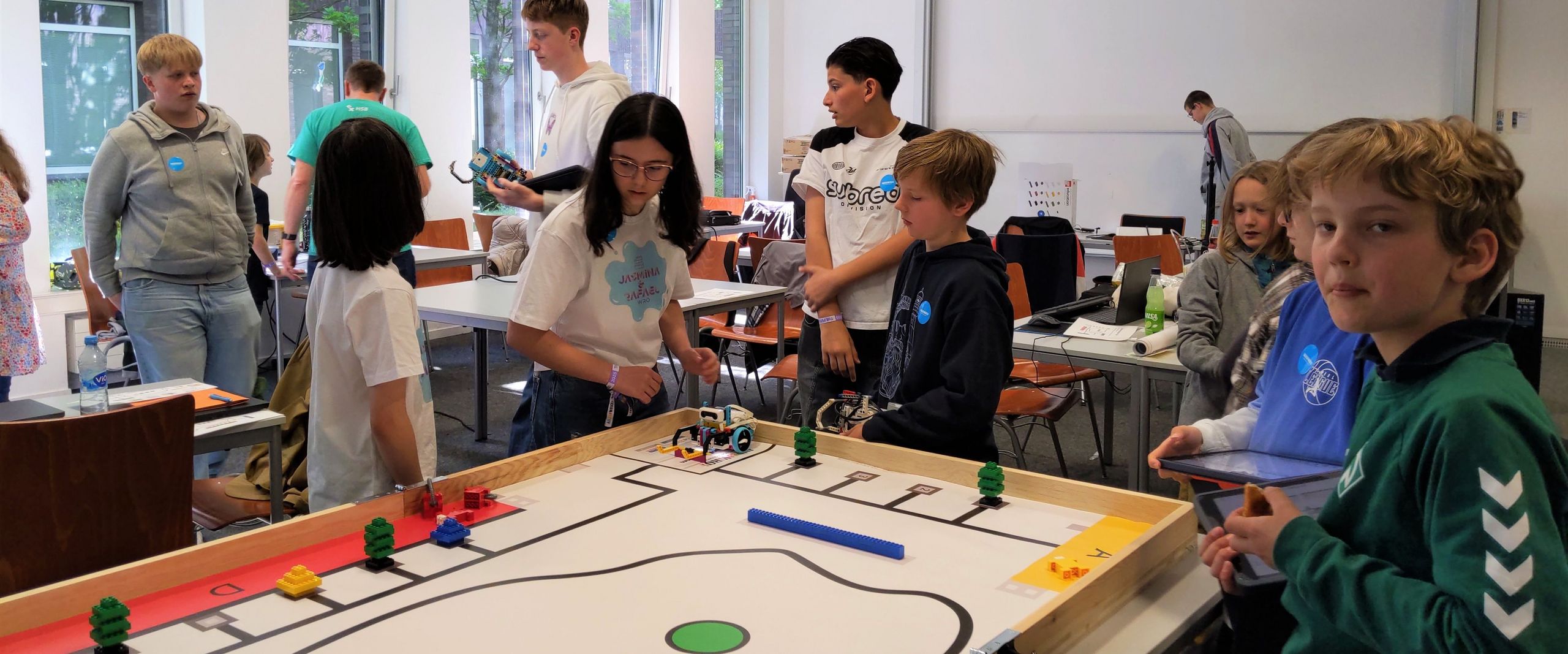 Mädchen und Jungen stehen um ein Spielfeld, auf dem kleine Roboter und Figuren fahren