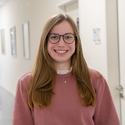 Auf dem Bild ist eine Studentin zusehen. Sie steht in einem wei&szlig;en Flur. Sie hat langes blondes Haar. Sie tr&auml;gt eine Brille und einen rosa f&auml;rbenden Pullover.