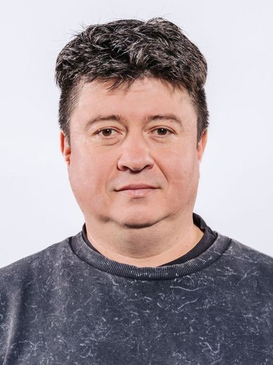 Auf dem Bild ist Kamil Iusupov zu sehen. er hat kurzes schwarzes Haar und trägt einen blau-grauen Pullover. 