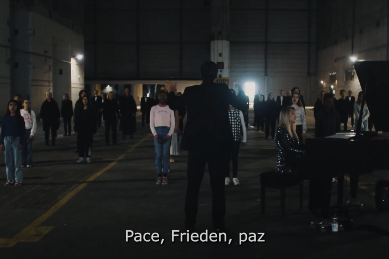 Videoausschnitt Song for Europe Gruppe von Menschen steht in einer dunklen Halle mit einem Klavierflügel