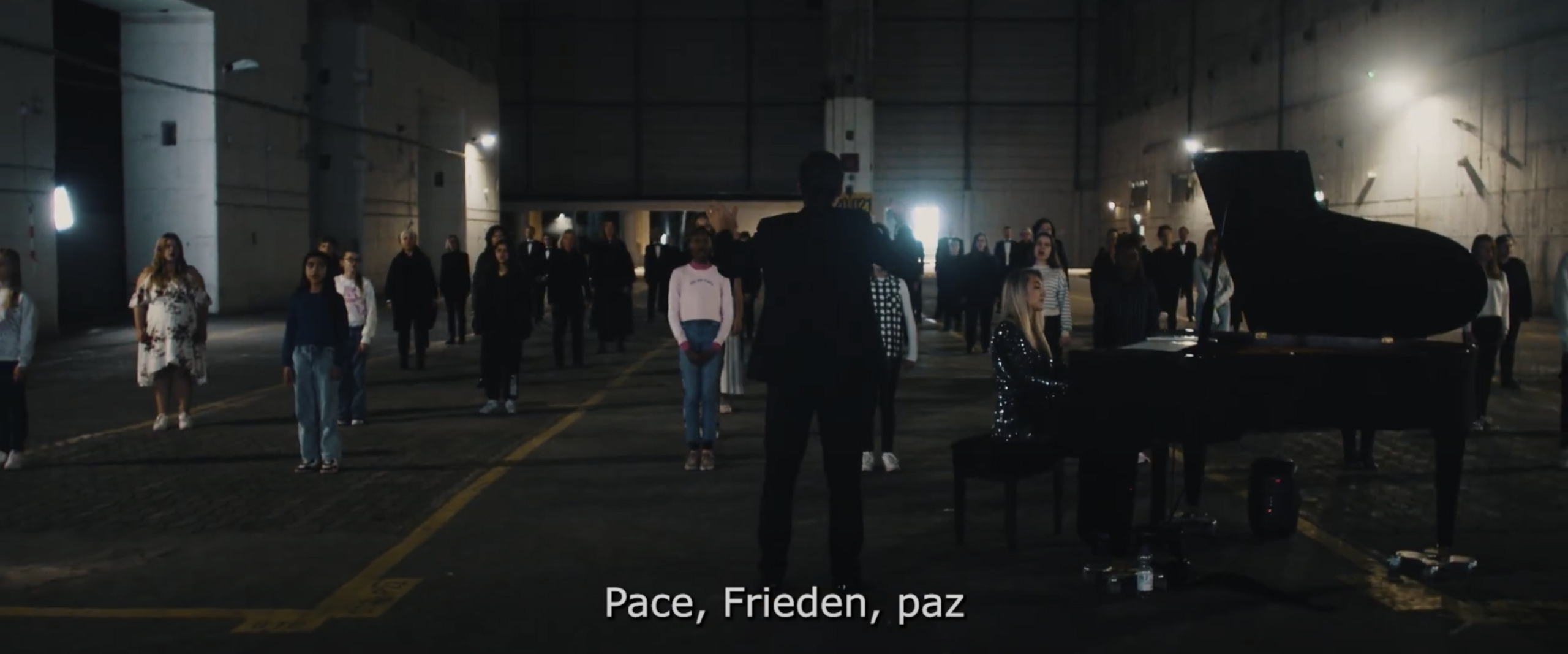 Videoausschnitt Song for Europe Gruppe von Menschen steht in einer dunklen Halle mit einem Klavierflügel