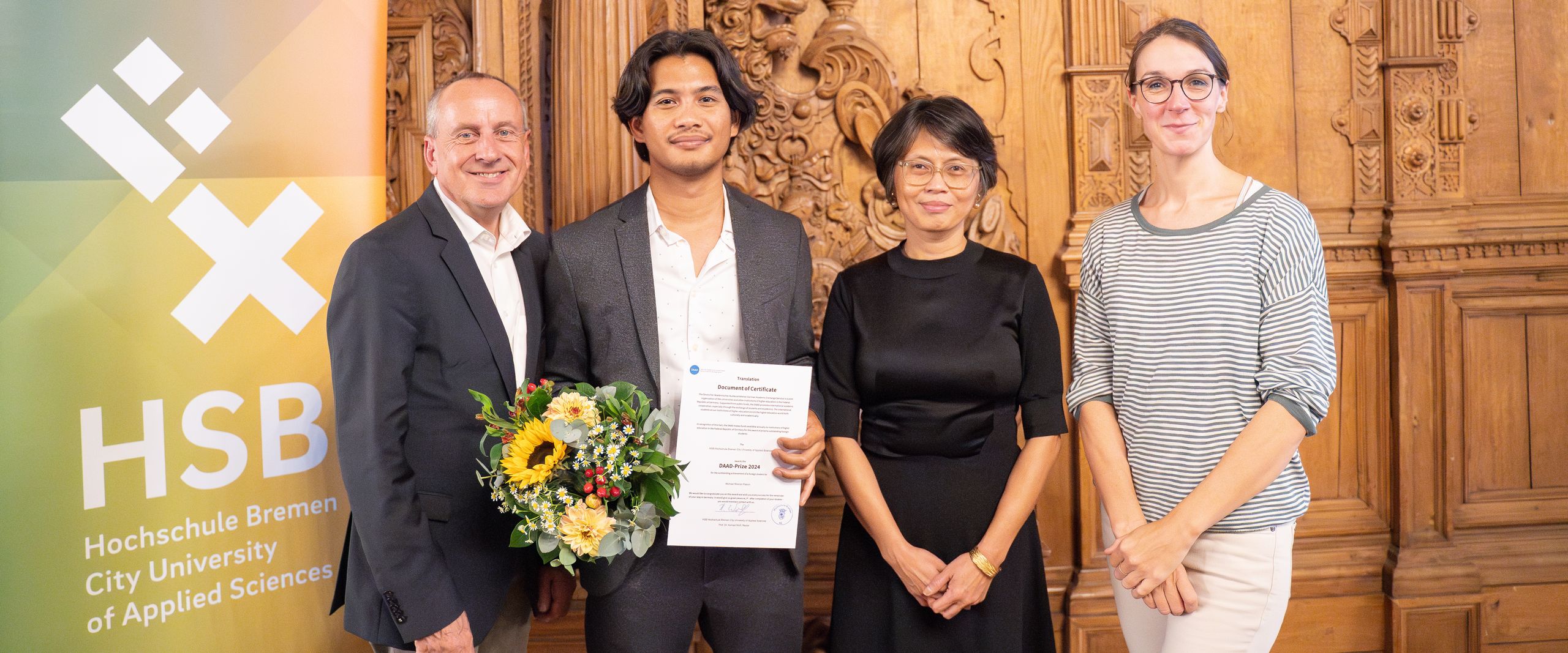 DAAD award winners with members of the University Management and his professor The photo shows four people looking into the camera. One is holding flowers and a certificate
