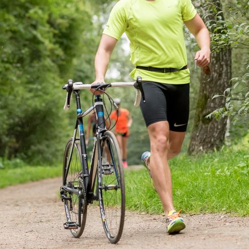 Ein Sportler l&auml;uft durch einen Wald und schiebt dabei ein Rennrad.