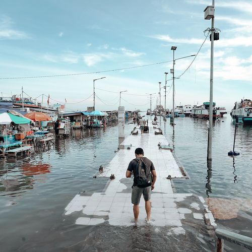 Ein Mensch watet &uuml;ber einen &uuml;berfuteten Steg. Links und rechts sind Wasser, Verkauffst&auml;nde und Boote.