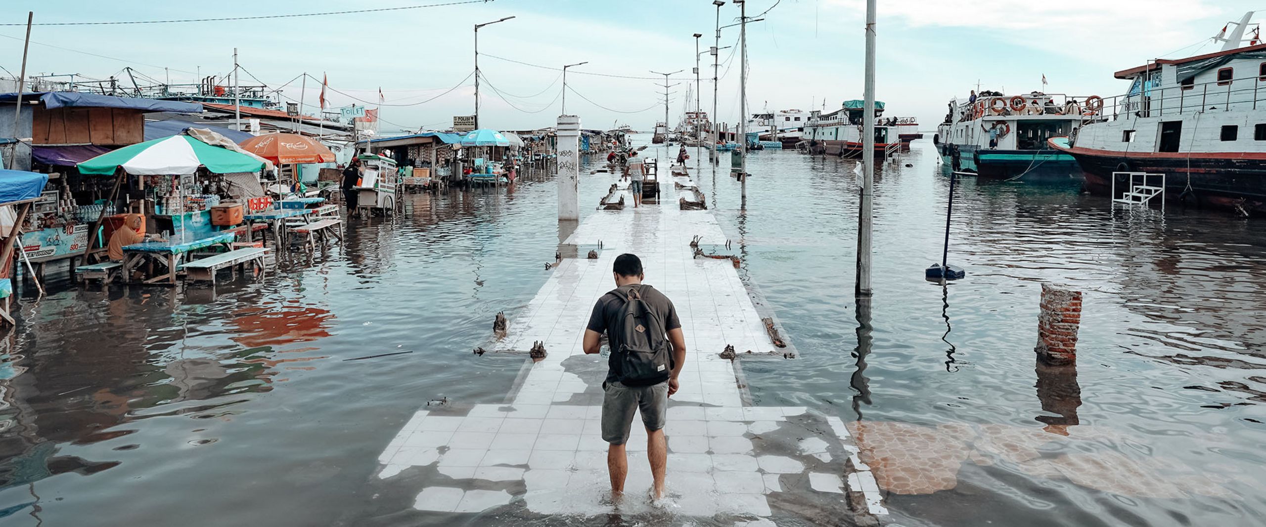 Ein Mensch watet über einen überfuteten Steg. Links und rechts sind Wasser, Verkauffstände und Boote.
