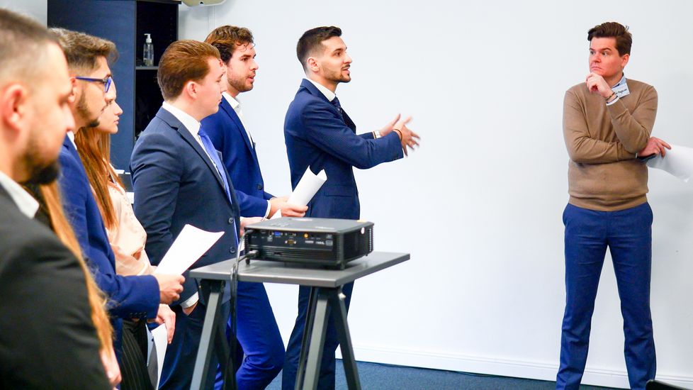 Mehrere Studierende stehen nebeneinander. Sie tragen in Business-Kleidung und halten teilweise Notizen in der Hand. Ein Student spricht und zeigt mit der Hand zur Seite. Ein Professor steht mit etwas Abstand seitlich von den Studierenden.