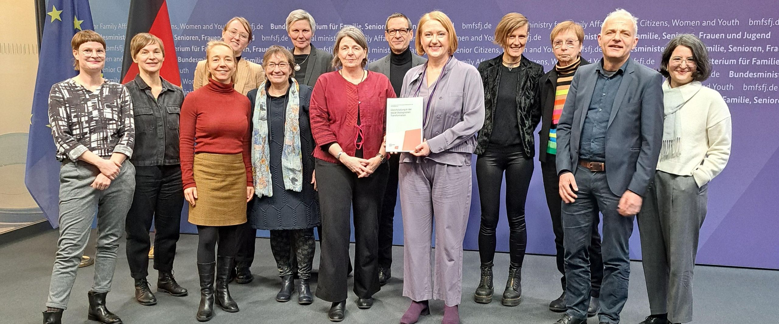 Auf dem Bild ist eine Gruppe von Leuten zusehen. Sie stehen in zwei Reihen und schauen in die Kamera. Eine Frau in der Mitte mit einem lila Hosenanzug hat einen ausgedruckten Bericht in der Hand. 