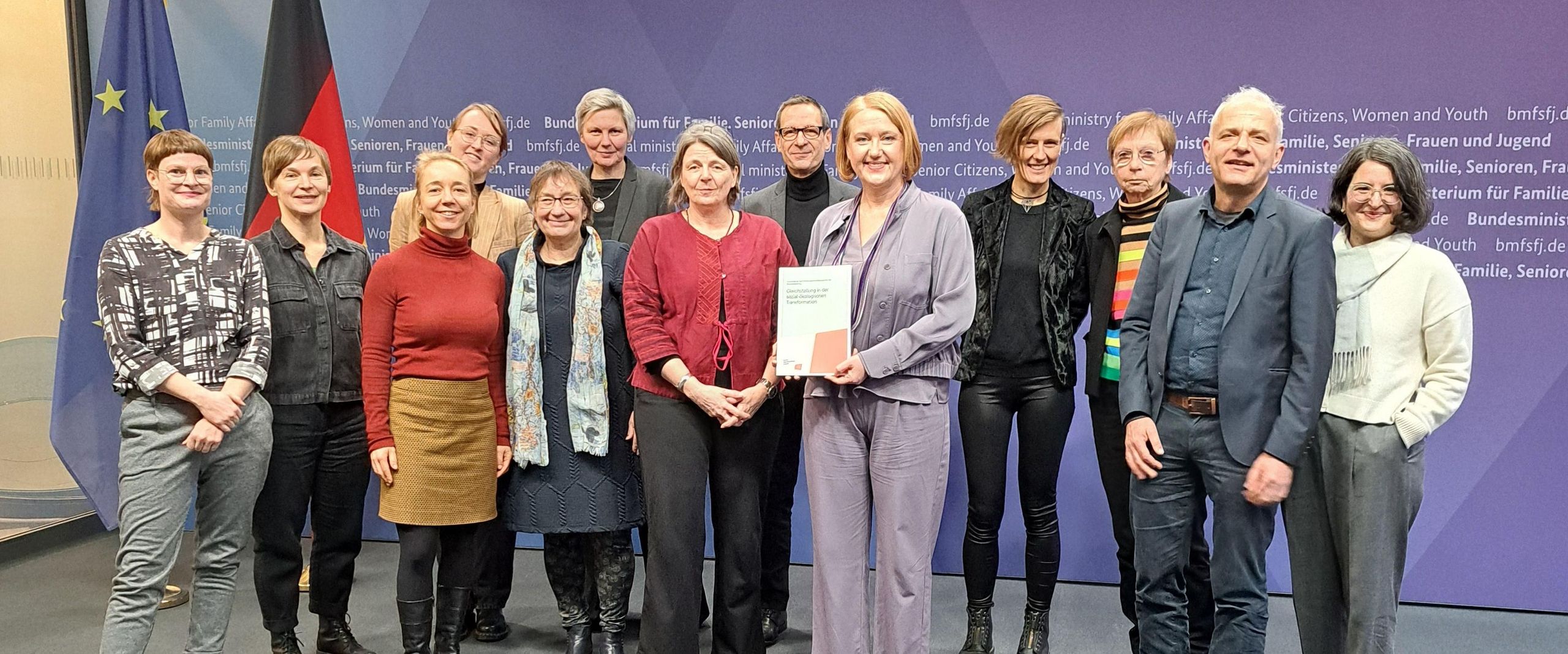 Auf dem Bild ist eine Gruppe von Leuten zusehen. Sie stehen in zwei Reihen und schauen in die Kamera. Eine Frau in der Mitte mit einem lila Hosenanzug hat einen ausgedruckten Bericht in der Hand. 
