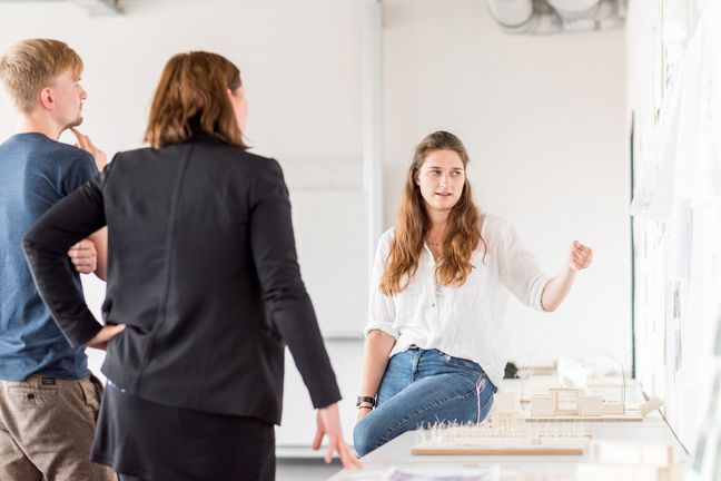 Eine Studentin präsentiert Prof. Andrea Dung und einem Kommilitonen ihre architektonischen Entwürfe.