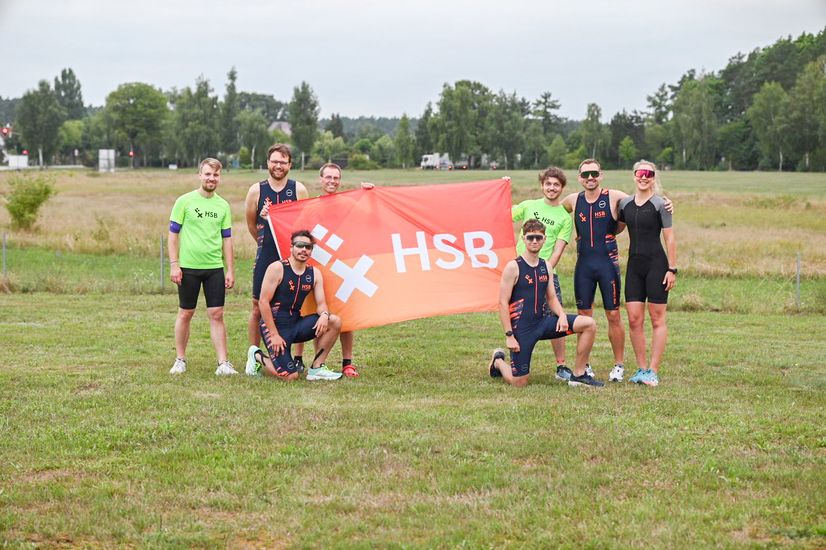 Das Team der HSB TriRunners Müritz auf einem Gruppenfoto beim Triathlon 2025. Der Sportbeauftragte Ingo Heidrich hält gemeinsam mit den Athleten eine HSB-Fahne