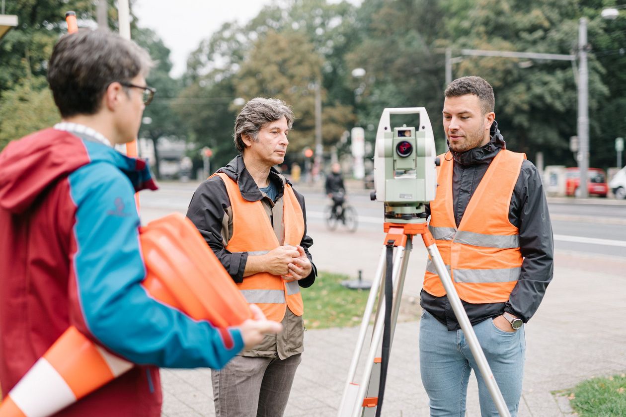 Ein Student in Warnweste bedient ein Messwerkzeug an der Stra&szlig;e, eine Dozent steht in Warnweste neben ihm, eine weitere Person steht daneben und h&auml;lt einen Stapel Pylone