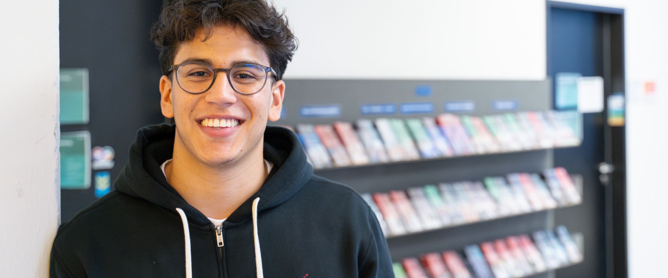 Nicolas Molina Sanchez Auf dem Bild ist Nicolas Molina Sanchez zusehen. Er steht mit der Schulter angelehnt an einer weißen Wand. Im Hintergrund sind bunte Flyer in einem Regal ausgestellt. Nicolas Sanchez trägt eine schwarze Sweatshirt-Jacke und hat braune lockige kurze Haare und eine runde Brille.