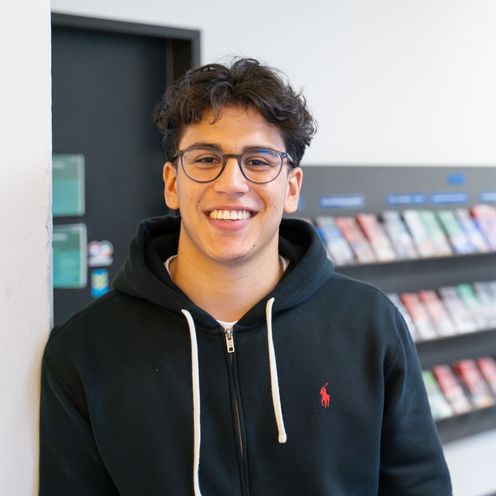 Auf dem Bild ist Nicolas Molina Sanchez zusehen. Er steht mit der Schulter angelehnt an einer wei&szlig;en Wand. Im Hintergrund sind bunte Flyer in einem Regal ausgestellt. Nicolas Sanchez tr&auml;gt eine schwarze Sweatshirt-Jacke und hat braune lockige kurze Haare und eine runde Brille. 