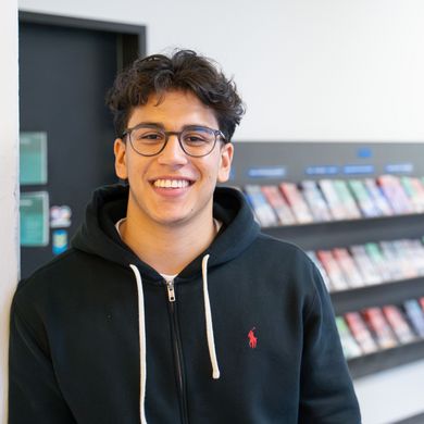 Auf dem Bild ist Nicolas Molina Sanchez zusehen. Er steht mit der Schulter angelehnt an einer weißen Wand. Im Hintergrund sind bunte Flyer in einem Regal ausgestellt. Nicolas Sanchez trägt eine schwarze Sweatshirt-Jacke und hat braune lockige kurze Haare und eine runde Brille. 