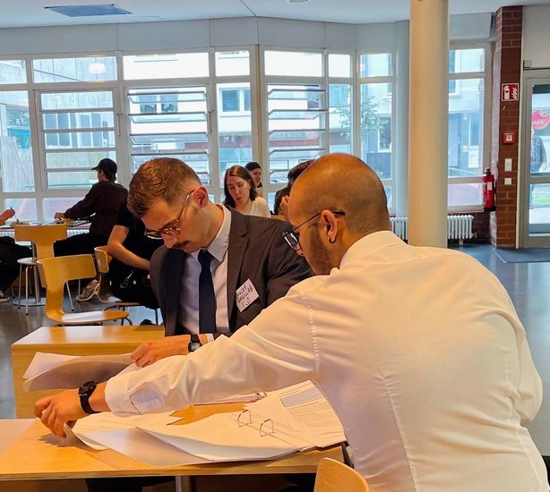 Auf dem Foto sind zwei Studierende zu sehen, die innerhalb eines simulierten Planspiels miteinander diskutieren und Unterlagen durch sehen. 