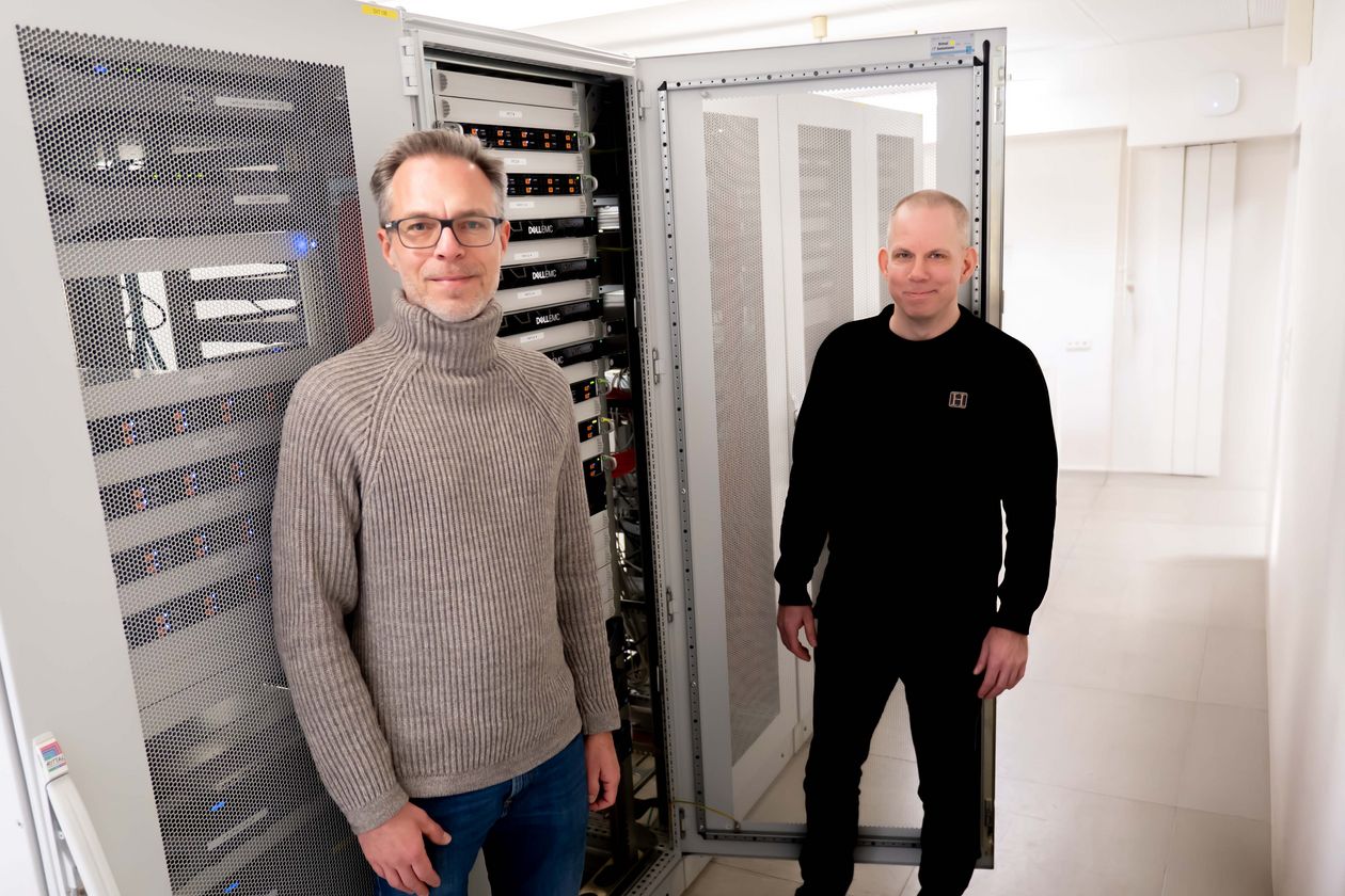 Zwei Männer stehen vor einem Server. Der Hintergrund ist hell und weiß.