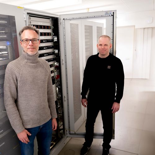 Zwei Männer stehen vor einem Server. Der Hintergrund ist hell und weiß.