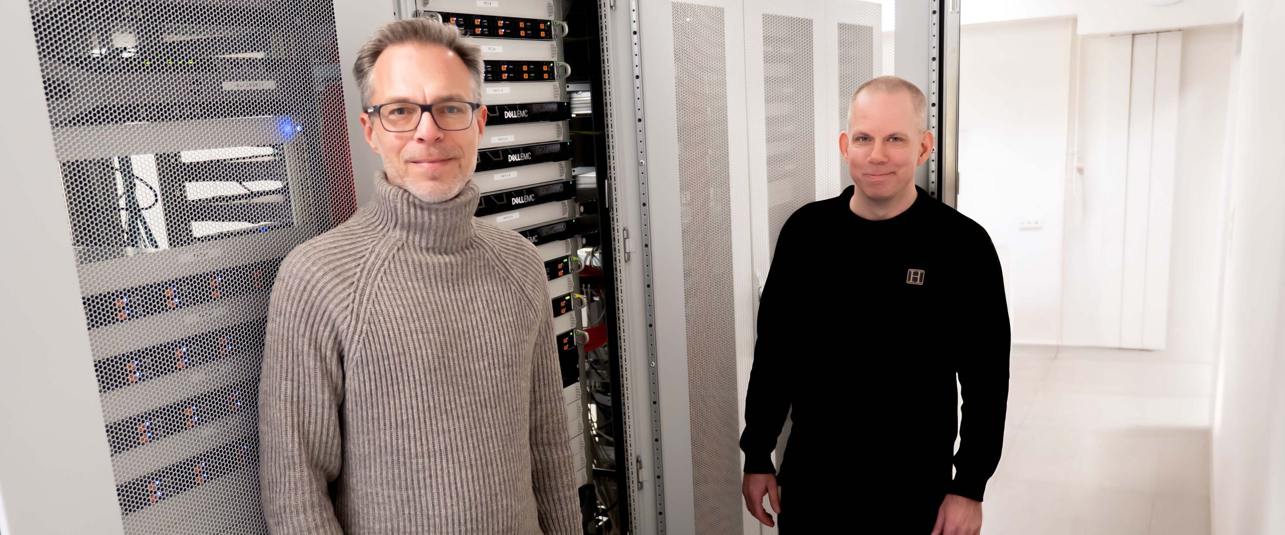 Zwei Männer stehen vor einem Server. Der Hintergrund ist hell und weiß.