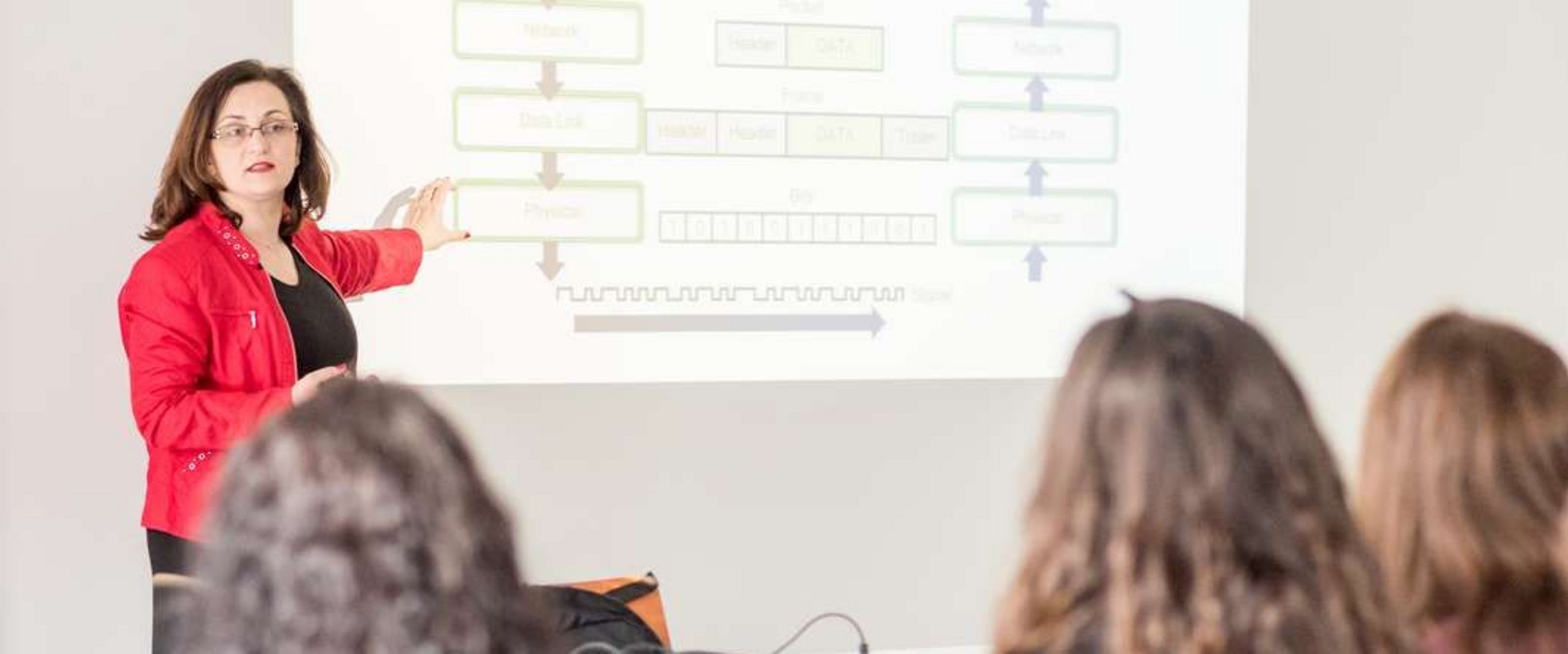 Professor Jasminka Matevska steht vor Studentinnen in einem Seminarraum. Im Hintergrund ist eine Grafik zu sehen, die von einem Beamer an die Wand projiziert wird.