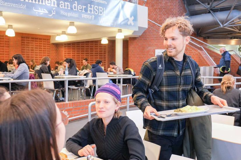 Mensa Essen Auf dem Bils sitzt eine Gruppe Studierenden an einem Tisch in der Mensa. Ein Studierender trägt gerade ein Tablett mit Essen zum Tisch.