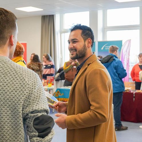 Auf dem Bild sind zwei Herren zusehen, die sich unterhalten. Der eine steht mit dem R&uuml;cken zur Kamera. In dem Hintergrund sind bunte St&auml;nde von Praxispartnern der HSB aufgebaut. 
