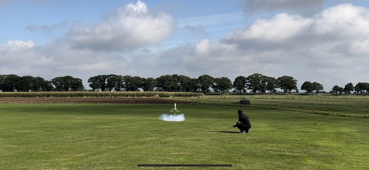 Eine grüße Wiese, auf der eine kleine Rakete startet. Ein Mensch sitzt mit Abstand davor.