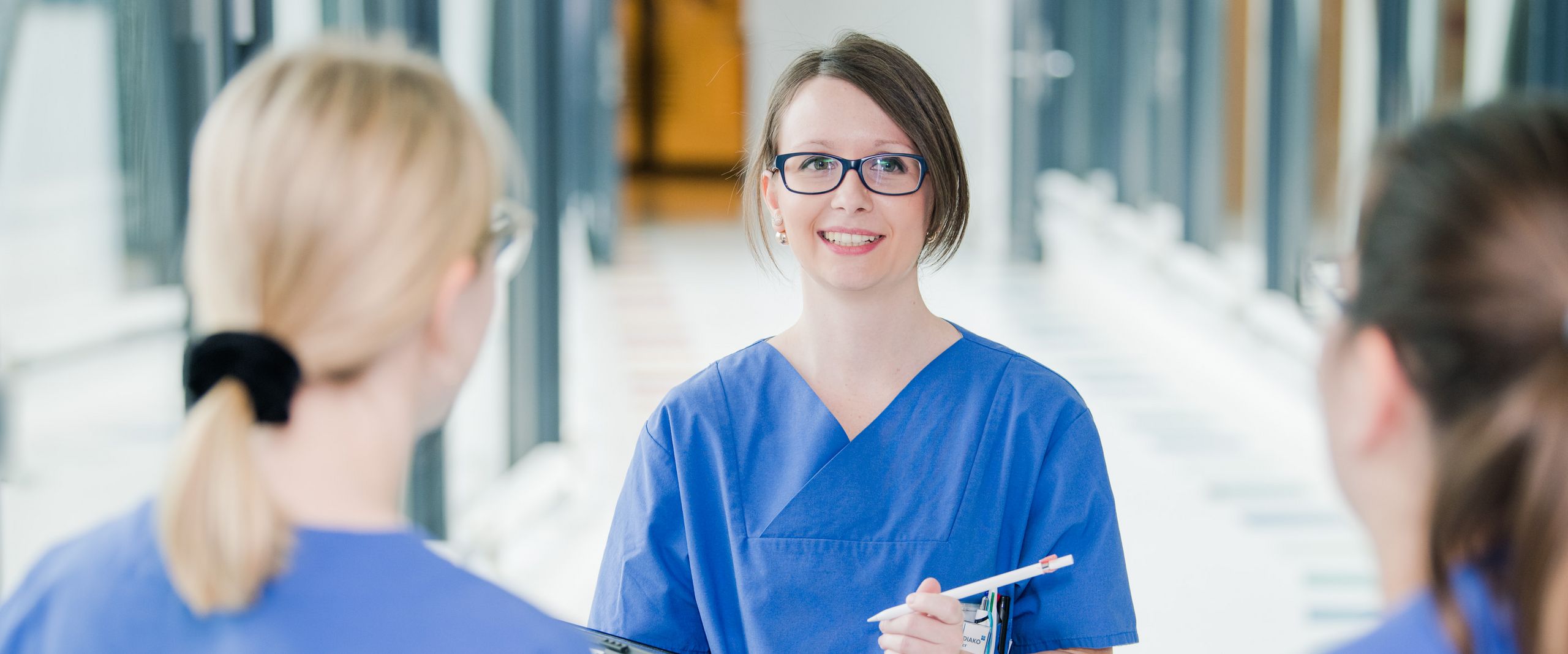 RS6769 Auf dem Bild sind drei Frauen in blauen Kasacks zu sehen. Zwei unscharf mit dem Rücken zur Kamera gedreht. Eine Person weiter hinten, schaut die beiden Frauen an und hat ein Tablet in der Hand. Auf sie ist der Fokus gerichtet.