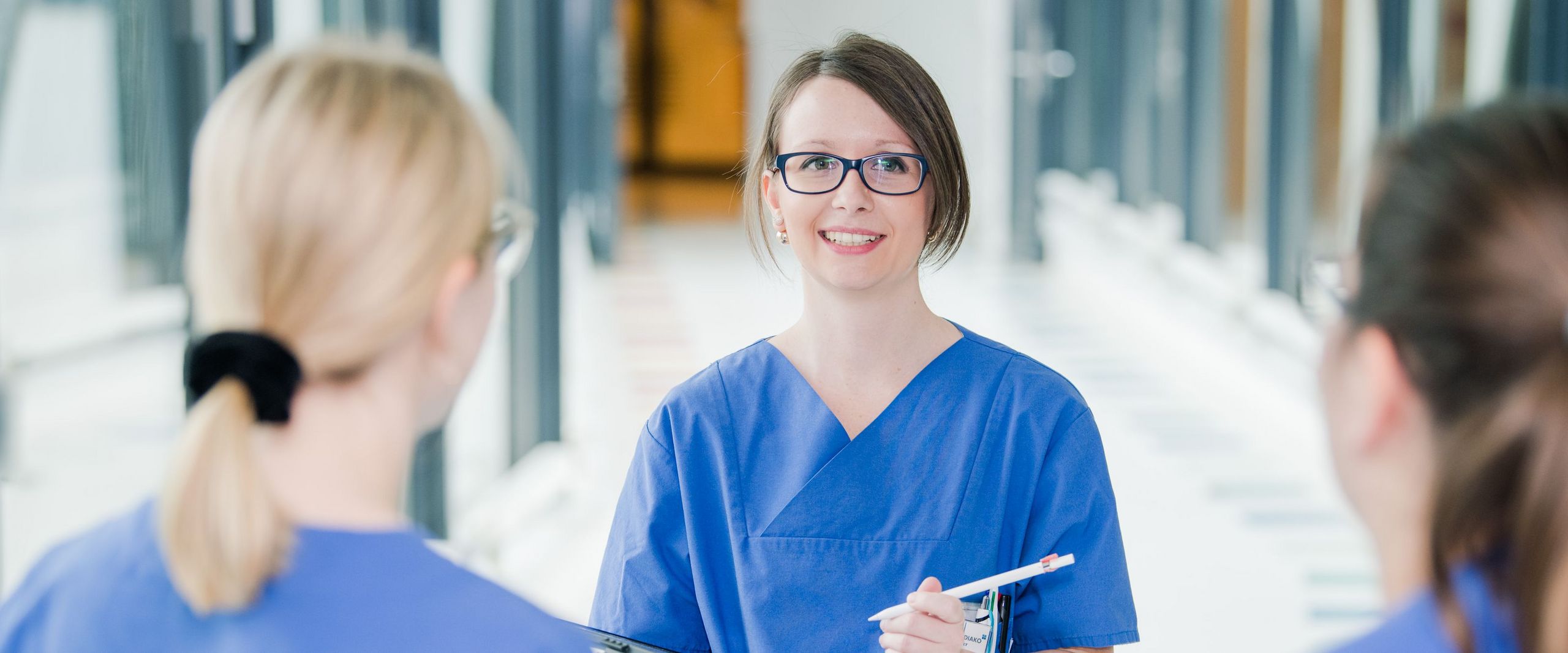 Auf dem Bild sind drei Frauen in blauen Kasacks zu sehen. Zwei unscharf mit dem Rücken zur Kamera gedreht. Eine Person weiter hinten, schaut die beiden Frauen an und hat ein Tablet in der Hand. Auf sie ist der Fokus gerichtet. 