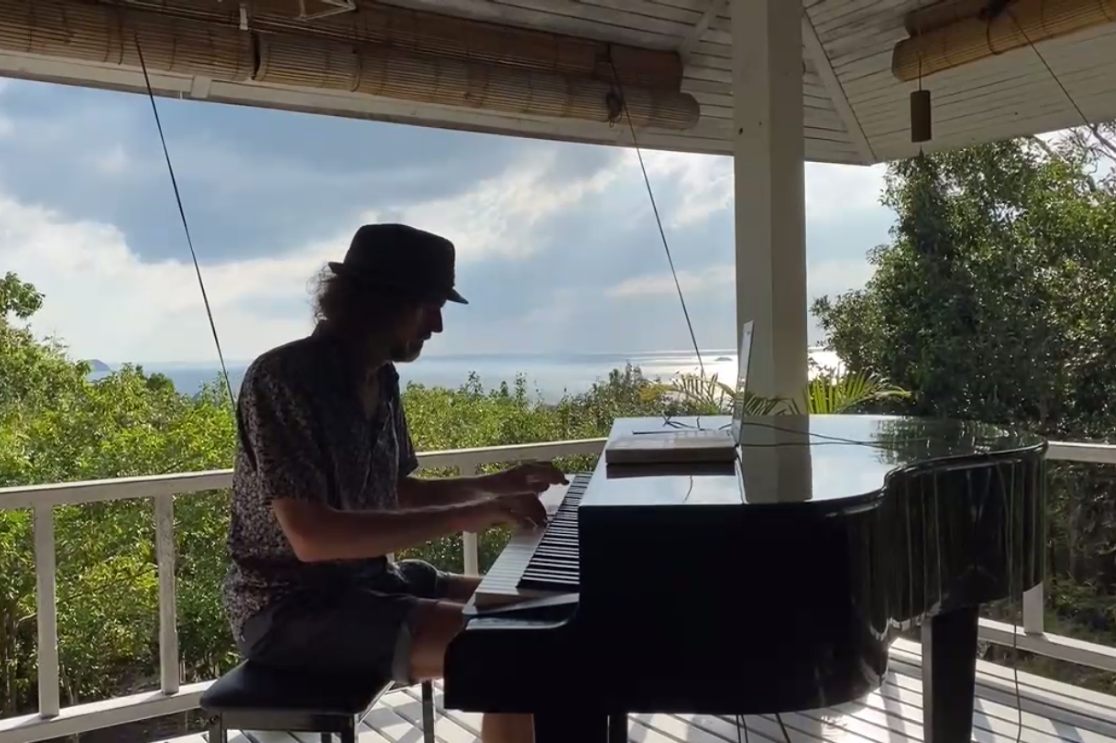 Ein Screenshot eines Zoom-Meetings auf dem ein Pianist zu sehen ist, der auf seinem Klavier auf einem Balkon ein Musikst&uuml;ck spielt. Im Hintergrund ist das Meer zu sehen. 