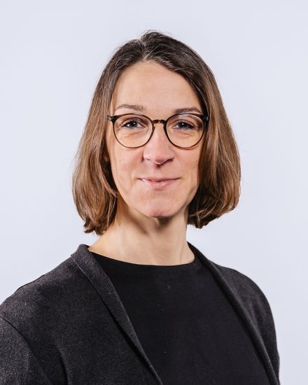 Auf dem Bild ist Annika Maschwitz zu sehen. Sie hat braunes, kinnlanges Haar. Sie trägt eine runde Brille und ein schwarzes T-Shirt unter einer dunkelgraunen Strickjacke.