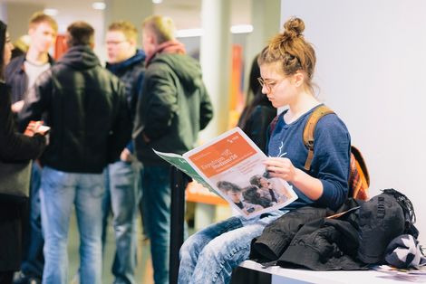Eine Besucherin des StudienINFOtags sitzt auf einem Tisch und liest im Programmheft des StudienINFOtags. Im Hintergrund ist verschwommen eine Gruppe von Besuchern im Gespräch zu sehen.