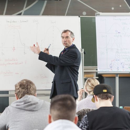 Professor Peter Schmidt steht vor einer Gruppe Studierender in einem Seminarraum und erkl&auml;rt etwas an einer wei&szlig;en Tafel.