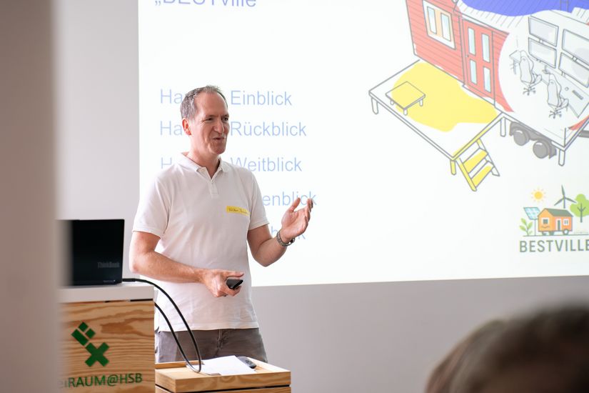 Auf dem Bild ist ein Mann zusehen, der einen Vortrag hält. Im Vordergrund ist unscharf das Publikum zu erkennen. 