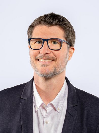 Auf dem Foto ist Sascha Behnk zusehen. Er hat kurze braune Haare. Er tr&auml;gt ein helles Hemd unter einem dunklen Sakko und eine Brille.