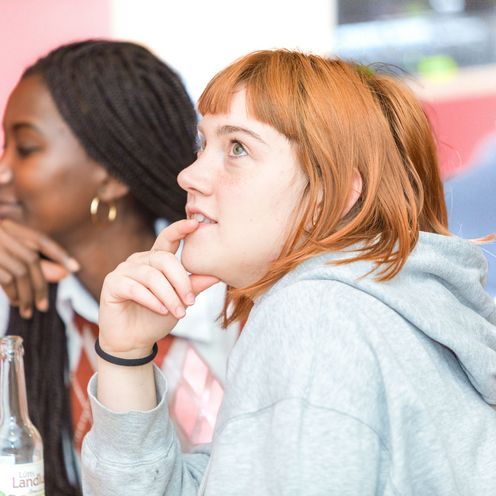 Zwei HSB Studierende seitlich ab Oberk&ouml;rper aufw&auml;rts fotografiert. Beide halten ihre H&auml;nde ans Kinn. Die vordere HSB Studierende blickt nach oben die hinter ihr sitzende blickt geradeaus. 