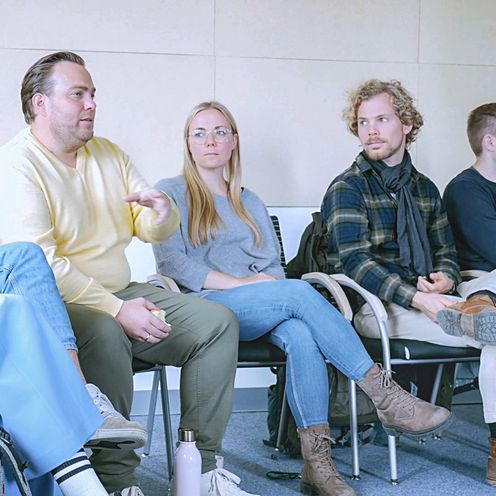 Bild mit M&auml;nnern und Frauen. Sie sitzen im Halbkreis zusammen und diskutieren.