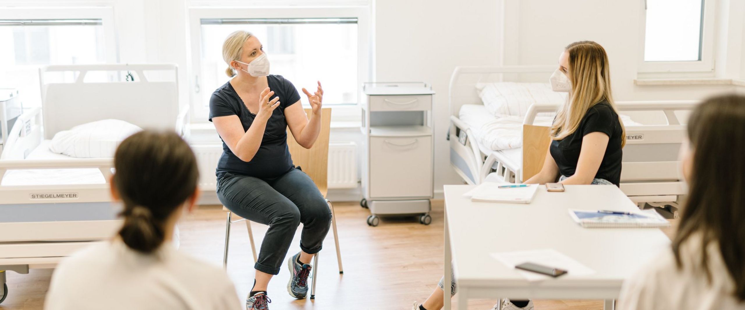 Eine Professorin sitzt neben einem Krankenbett und erklärt ihren Studierenden etwas.