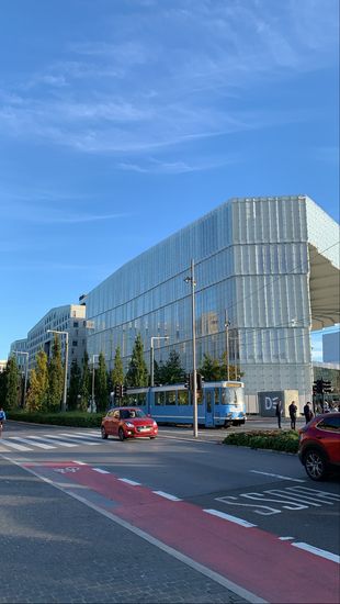 Auf diesem Bild ist die BI Norwegian Business University von außen zu sehen. Es ist ein sehr modernes, helles Gebäude mit vielen Fenstern. Vor dem Gebäude fahren Autos sowie eine Straßenbahn. Der blaue Himmer spielgelt sich in den Fenstern. 