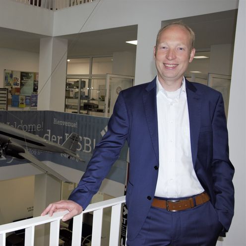 L&auml;chelnder Mann im blauen Anzug, der an einem wei&szlig;en Gel&auml;nder lehnt. Im Hintergrund sieht man ein Modell eines Traditionsfliegers.