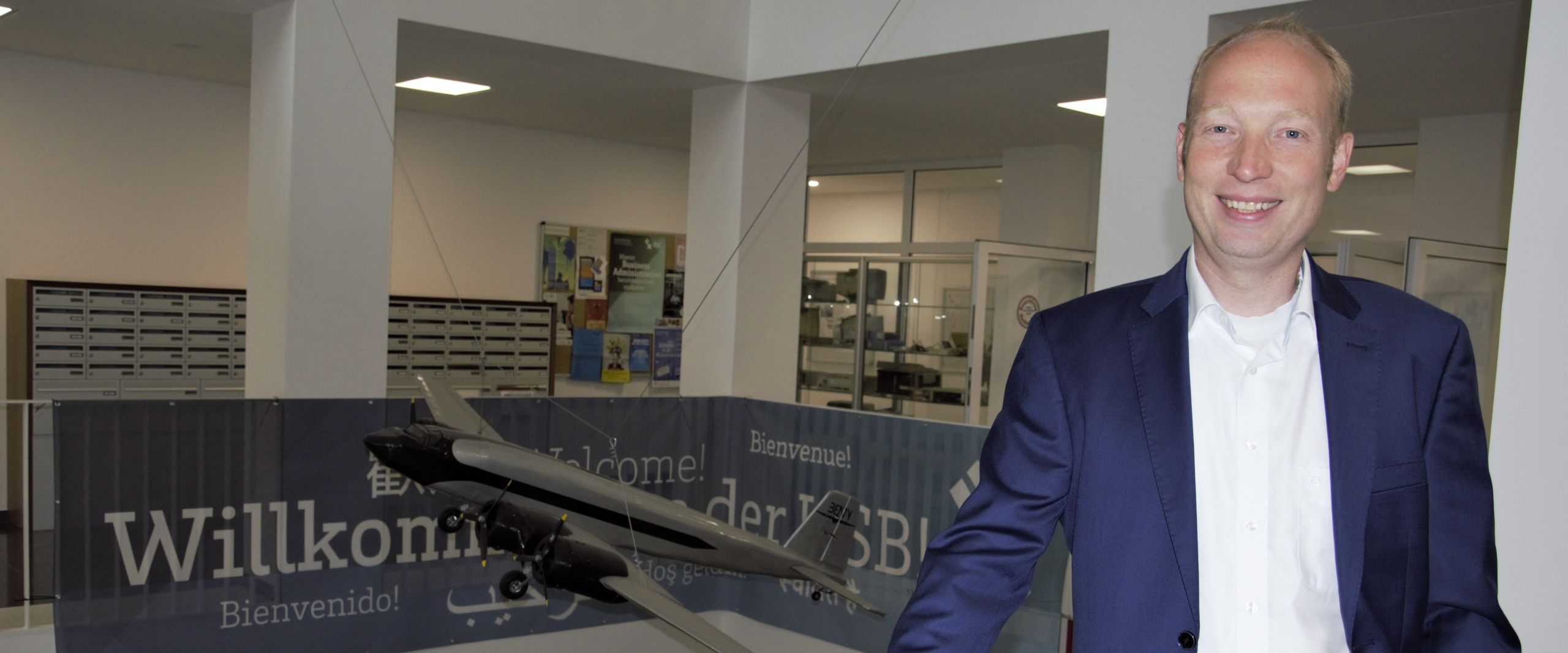 Jorgen von der Brelie im ZIMT Lächelnder Mann im blauen Anzug, der an einem weißen Geländer lehnt. Im Hintergrund sieht man ein Modell eines Traditionsfliegers.