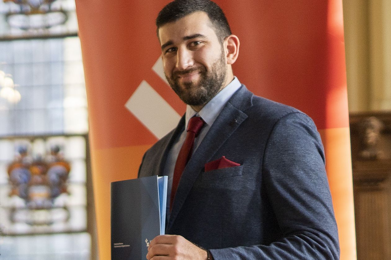 Mann mit Bart steht vor einem organgenen Rollup und hält eine blaue Mappe in der hand. Er lacht in die Kamera.