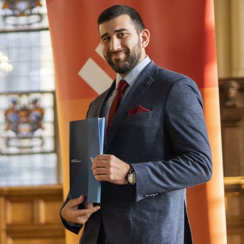 Mann mit Bart steht vor einem organgenen Rollup und h&auml;lt eine blaue Mappe in der hand. Er lacht in die Kamera.