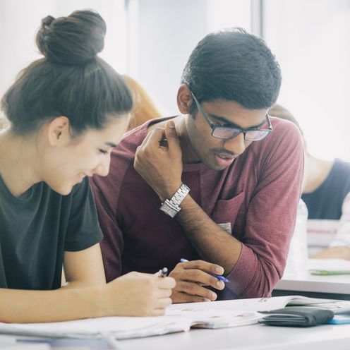 Eine Studentin und ein Student sitzen nebeneinander und blicken gemeinsam auf Unterlagen vor sich auf dem Tisch.