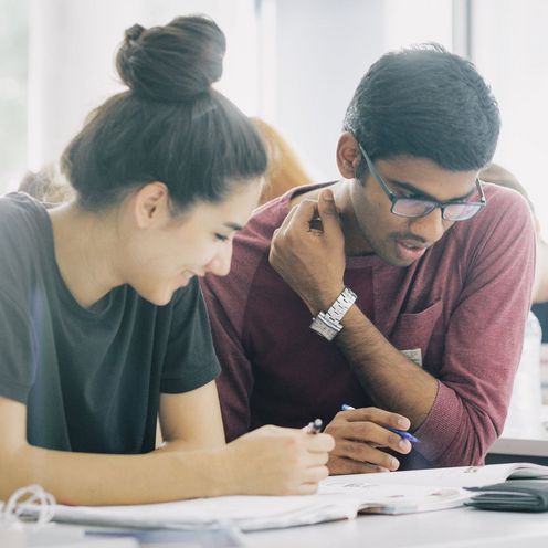 Eine Studentin und ein Student sitzen nebeneinander und blicken gemeinsam auf Unterlagen vor sich auf dem Tisch.